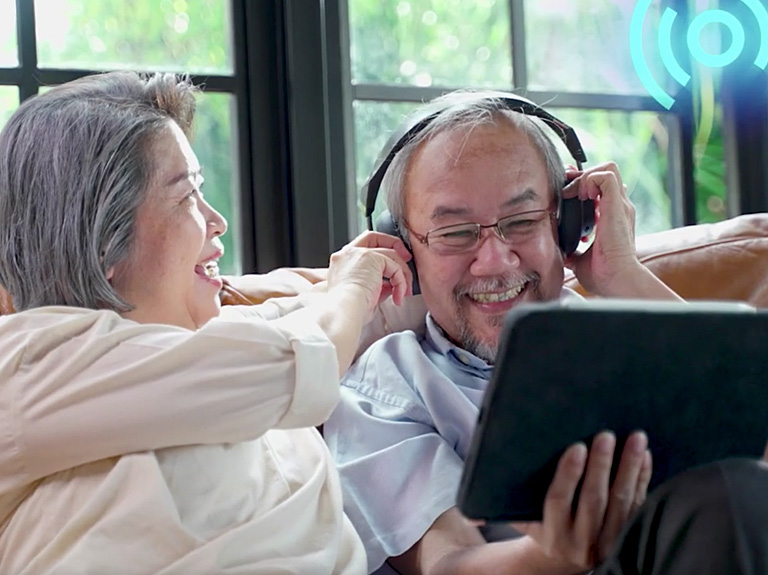 An older man and woman laugh while putting headphones on the man and watching a tablet screen