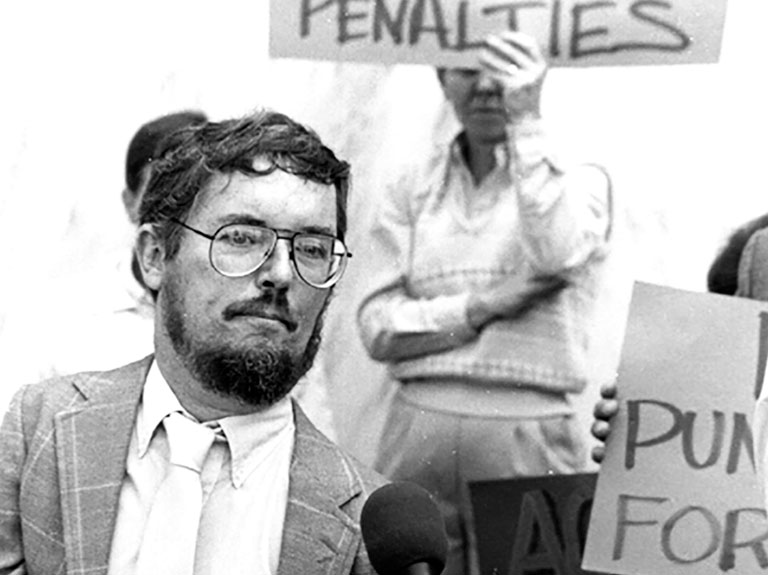 Historian Paul Longmore - Man dressed in suit and wearing glasses stands behind microphone at rally with people holding signs in the background